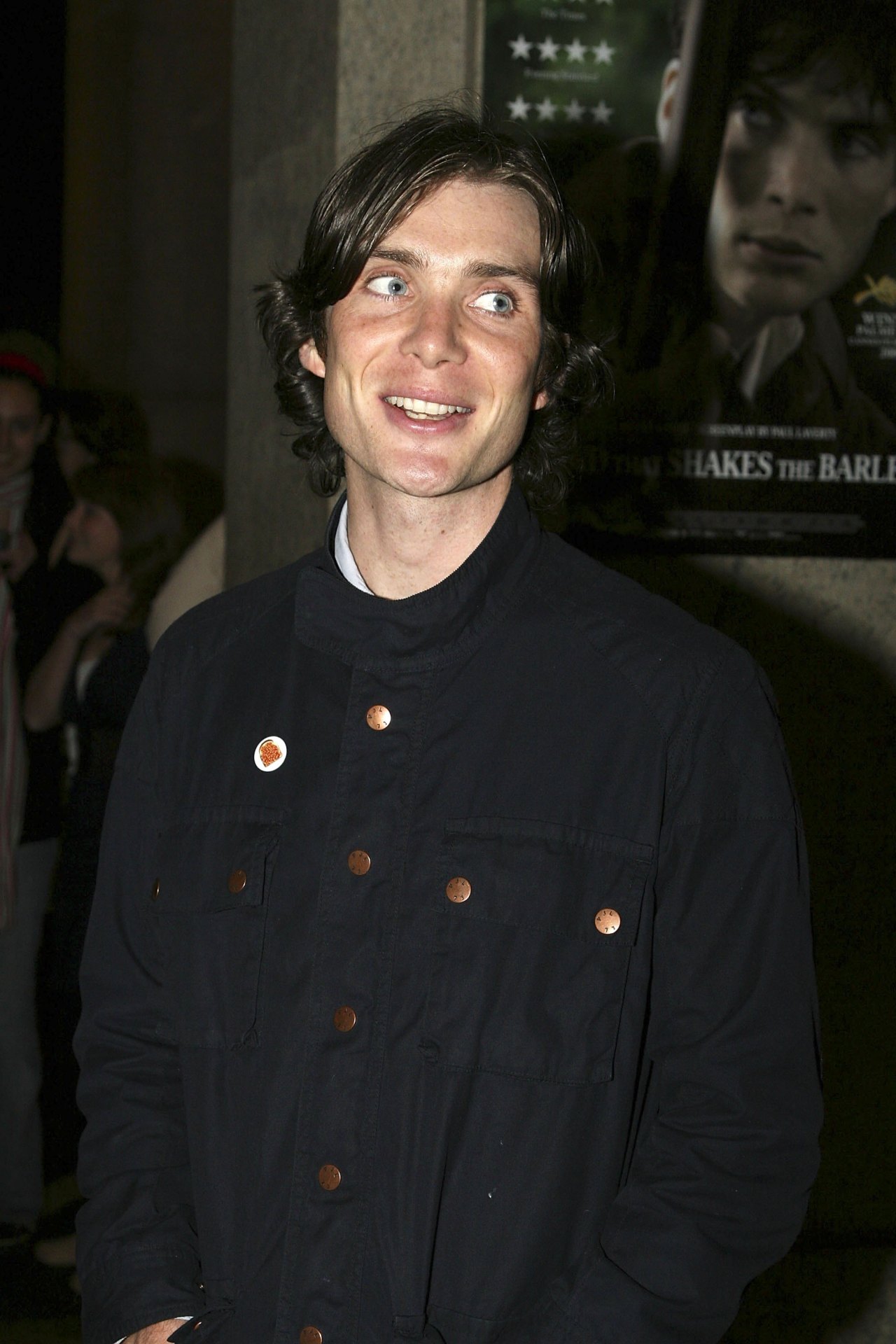 Cillian Murphy smiling with wide eyes at a film premiere, wearing a dark navy jacket with copper buttons and a pin badge.