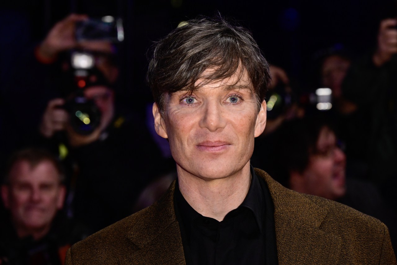 Cillian Murphy in a brown tweed blazer looking directly at the camera on a red carpet, photographers visible in the background.