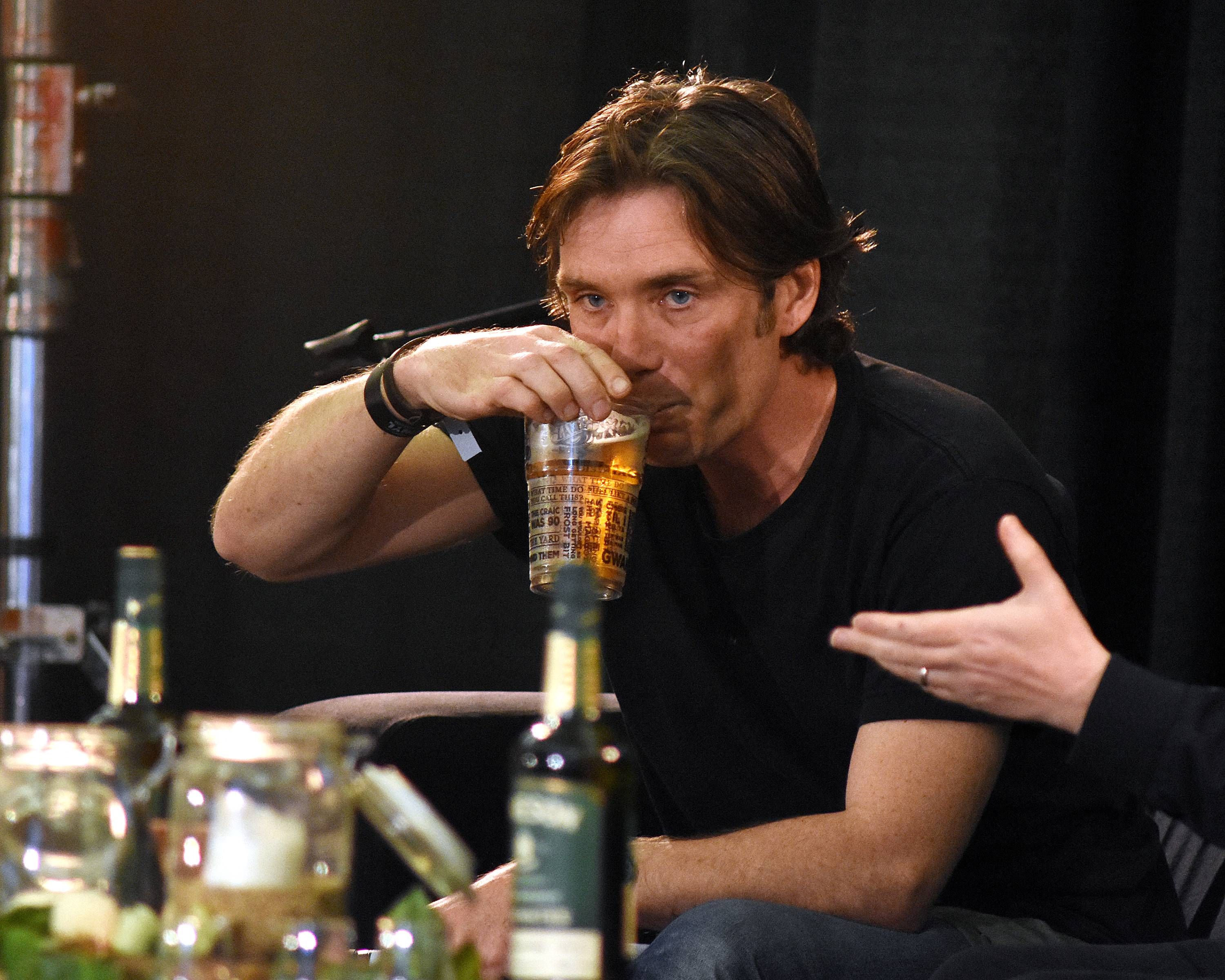 Click to open the image gallery Man with brown hair drinking a beer from a printed pint glass at an indoor festival event, bottles on the table in front of him.