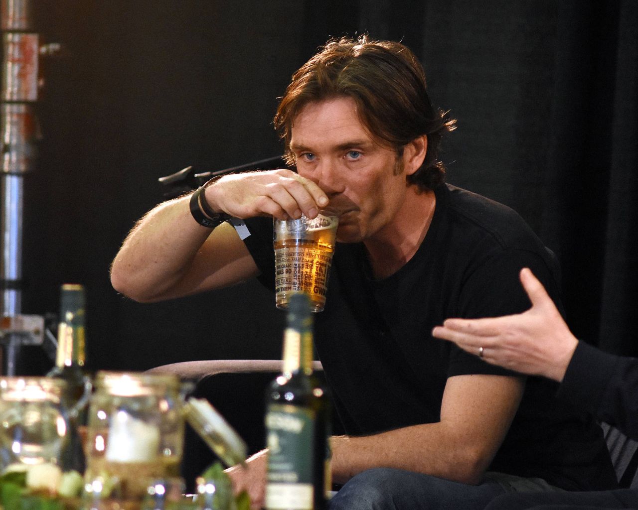Man with brown hair drinking a beer from a printed pint glass at an indoor festival event, bottles on the table in front of him.