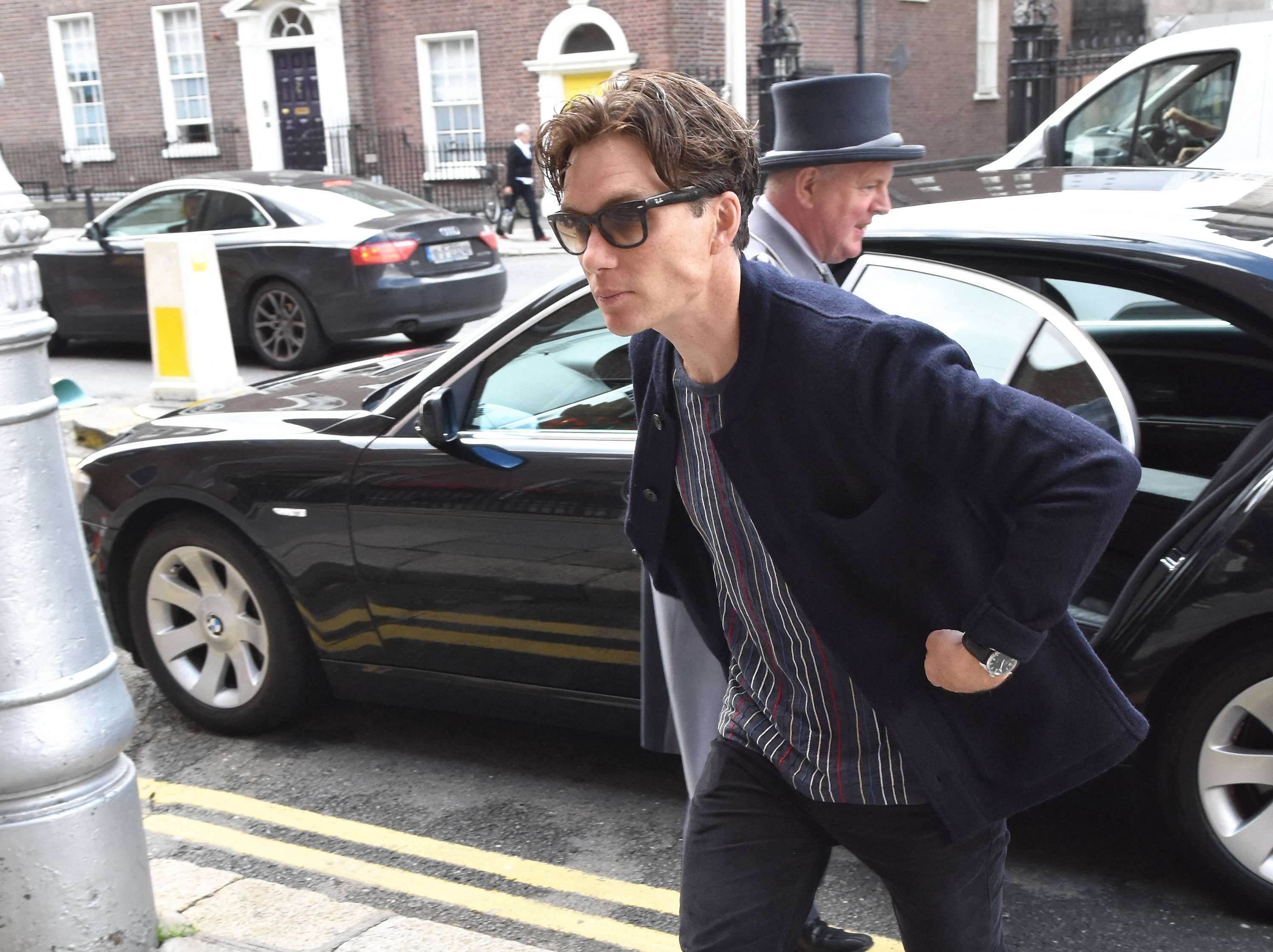Click to open the image gallery Man in sunglasses and a navy coat leaning forward while getting out of a car on a city street, with a man in a top hat visible behind him.