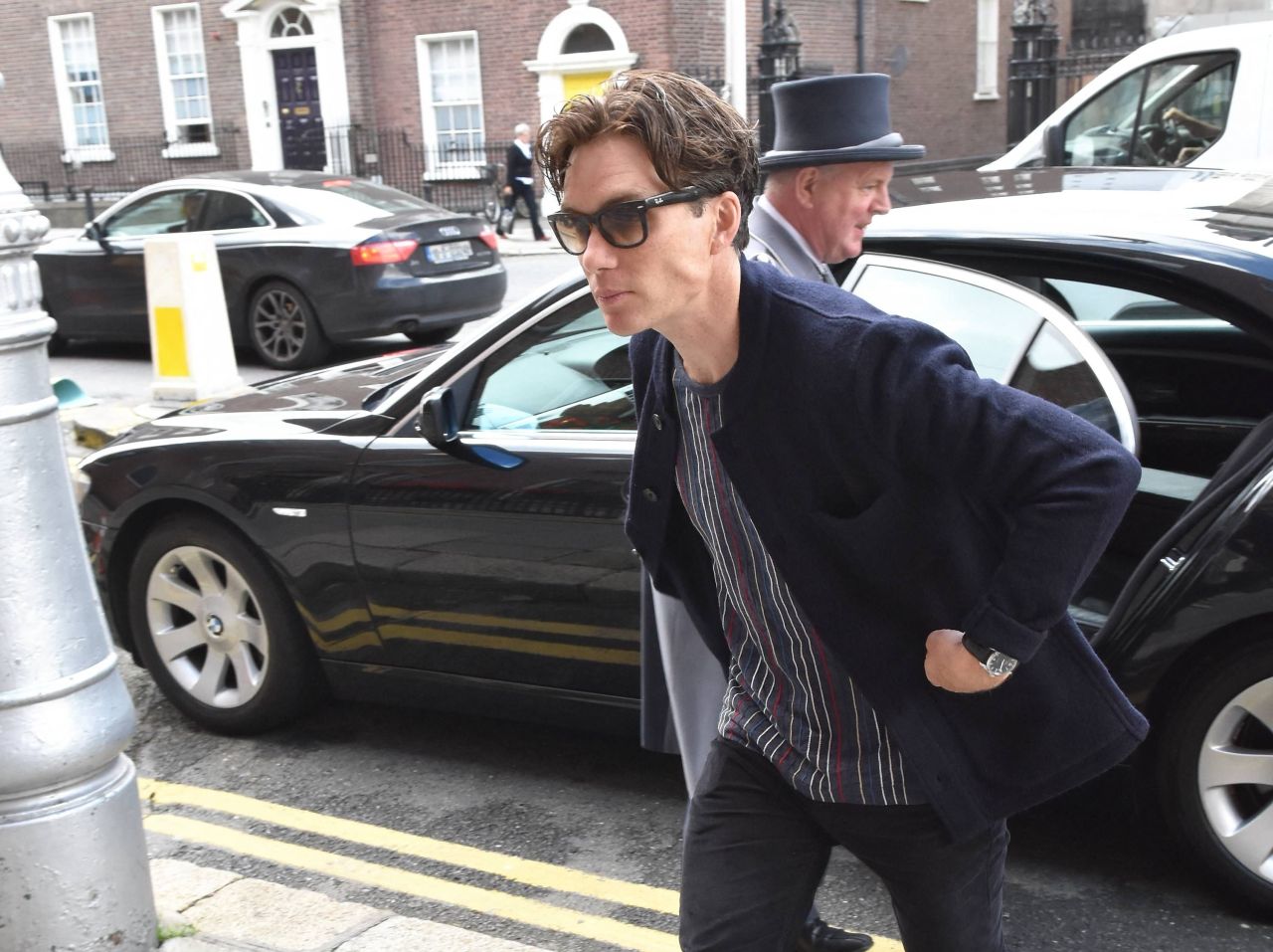Man in sunglasses and a navy coat leaning forward while getting out of a car on a city street, with a man in a top hat visible behind him.