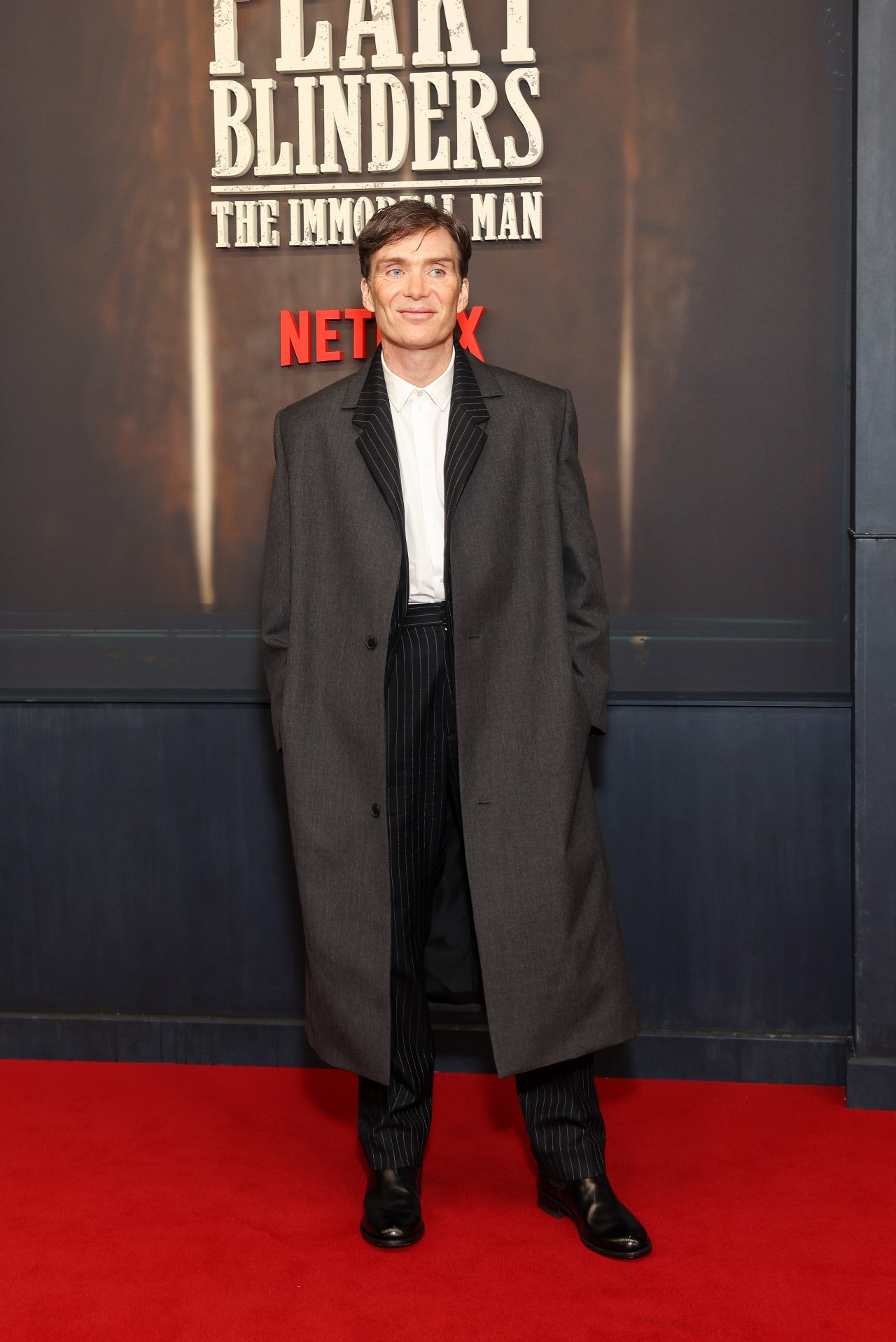 Man in a long grey overcoat and black pinstripe trousers posing on a red carpet in front of a Peaky Blinders: The Immortal Man Netflix backdrop.