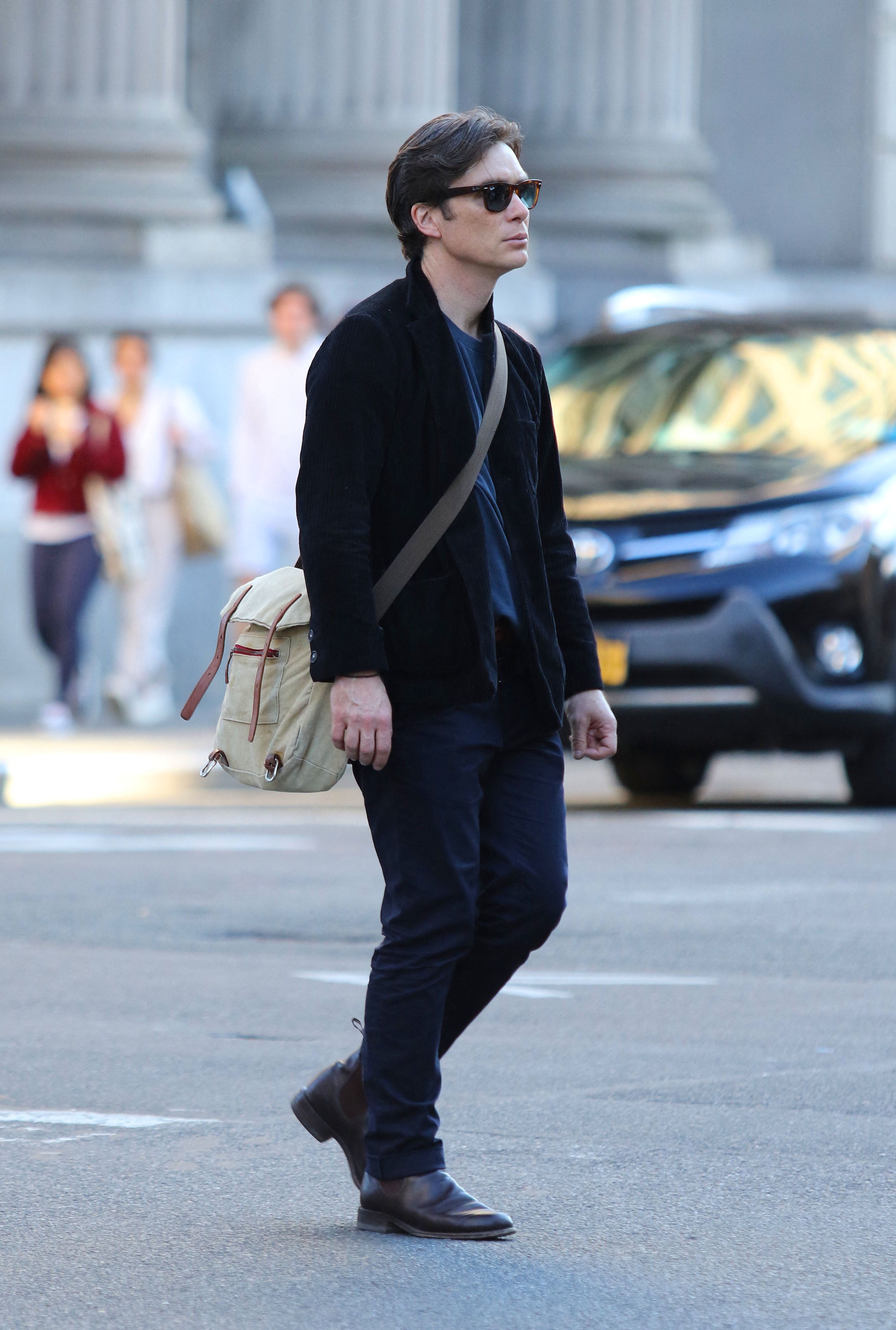 Click to open the image gallery Man in a black blazer, dark jeans, and sunglasses carrying a beige canvas backpack while crossing a city street.