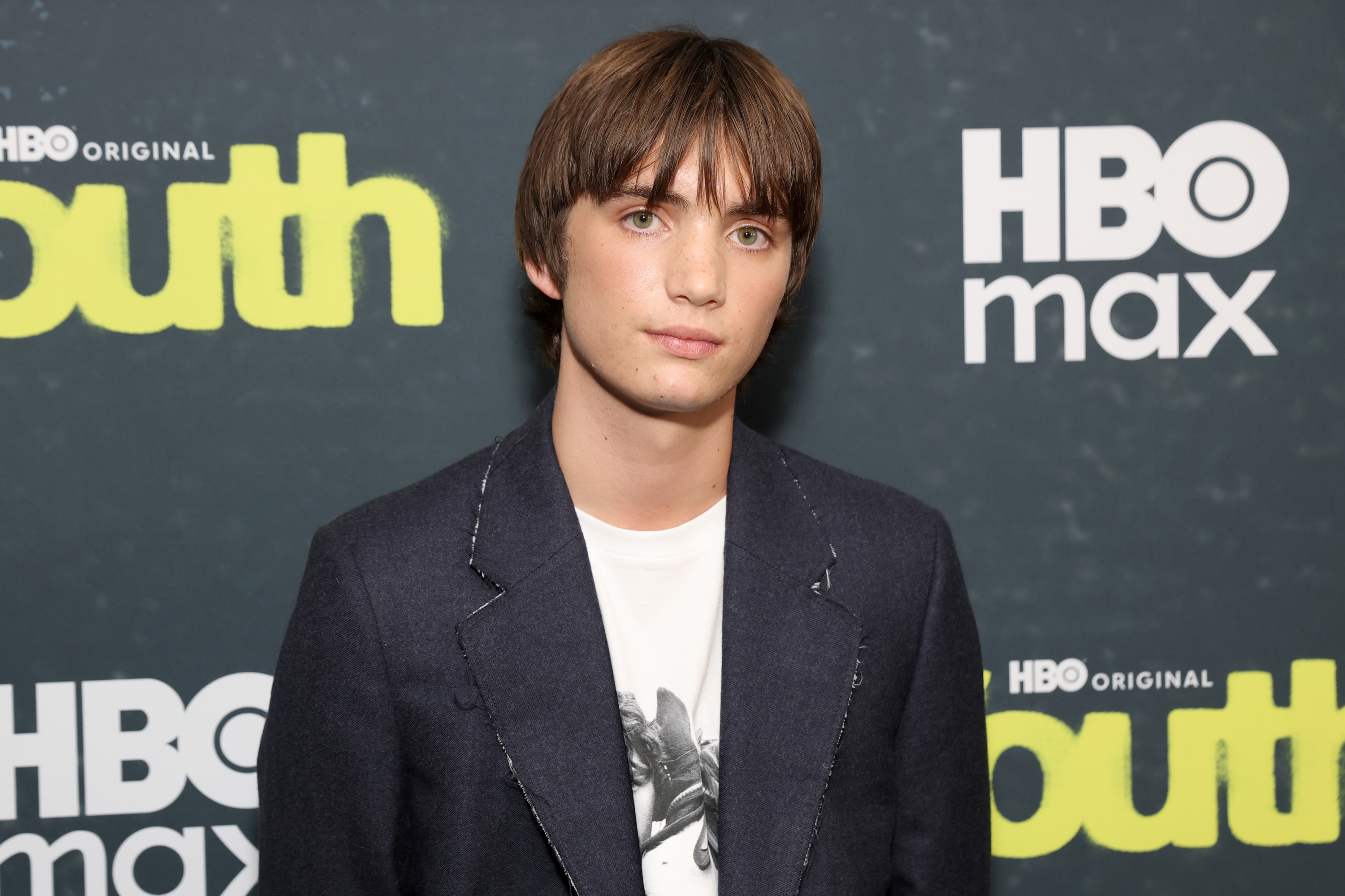 Click to open the image gallery Young man with brown bowl-cut hair and green eyes in a dark blazer over a graphic tee at an HBO Youth premiere backdrop.