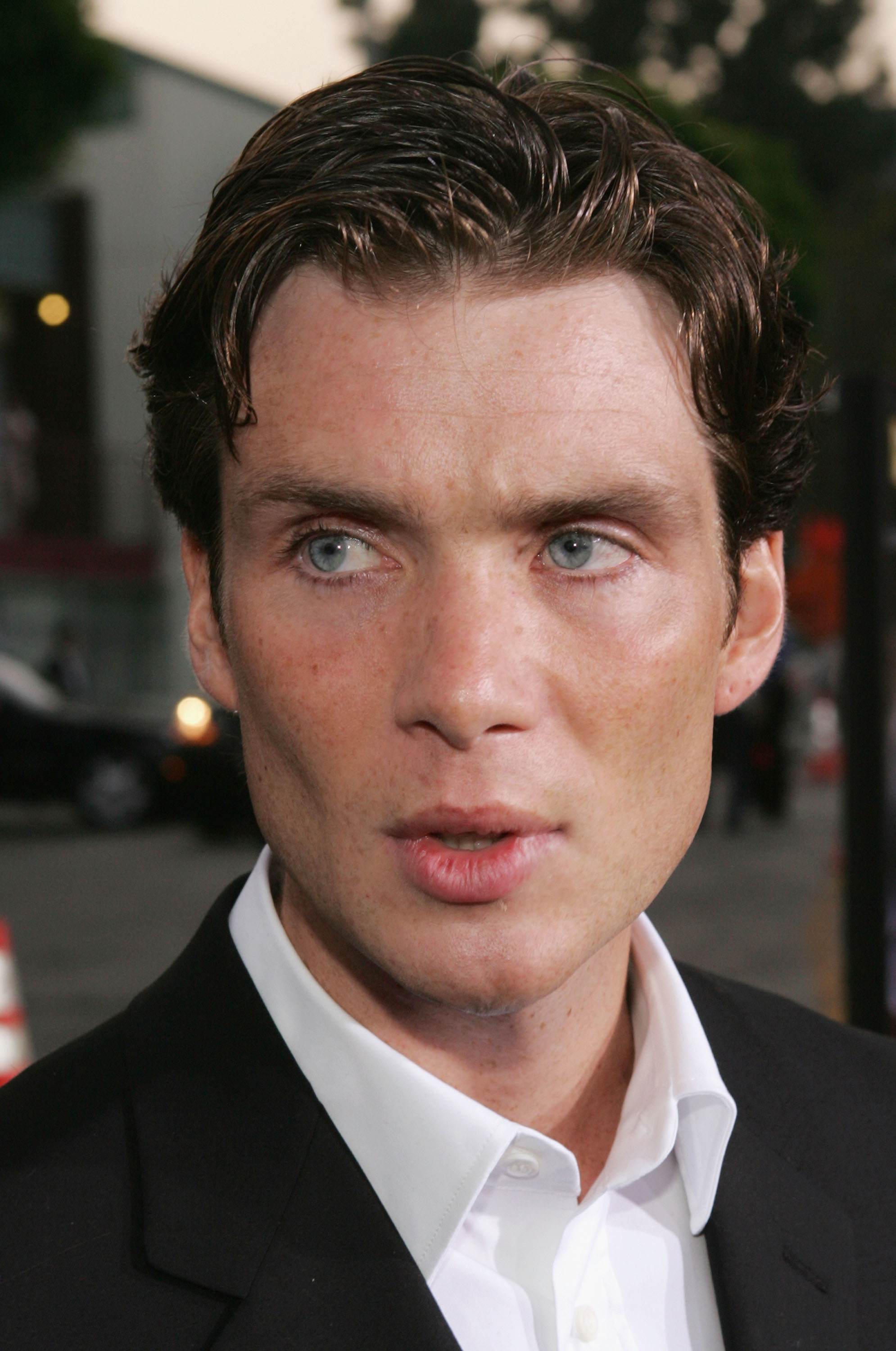 Click to open the image gallery Young man with slicked-back dark hair and blue eyes in a black blazer and white open-collar shirt at an outdoor film premiere.