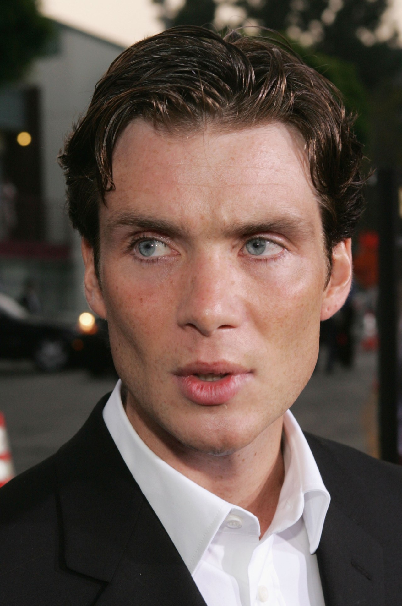 Young man with slicked-back dark hair and blue eyes in a black blazer and white open-collar shirt at an outdoor film premiere.