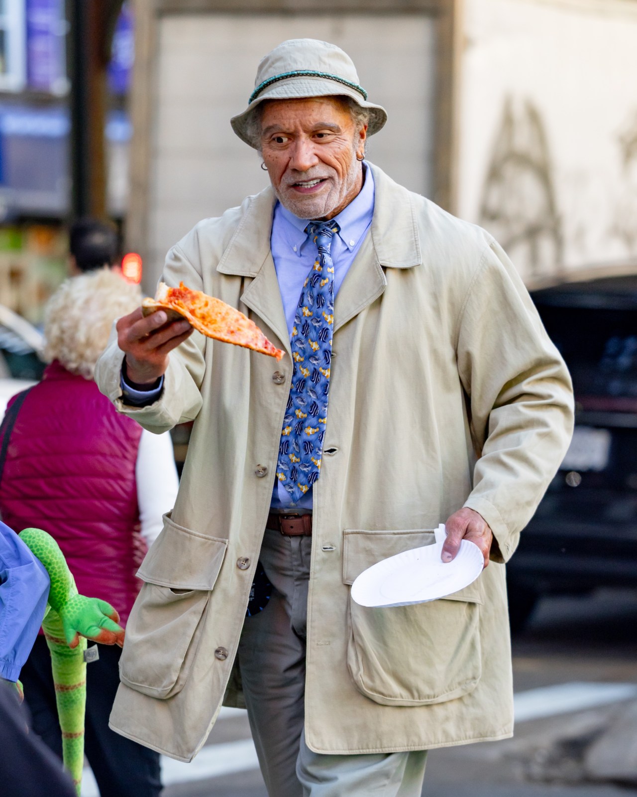 Dwayne Johnson in prosthetic makeup, gray curly hair, bucket hat, and tan overcoat walking alongside young co-star Theo Clark Leber on a film set