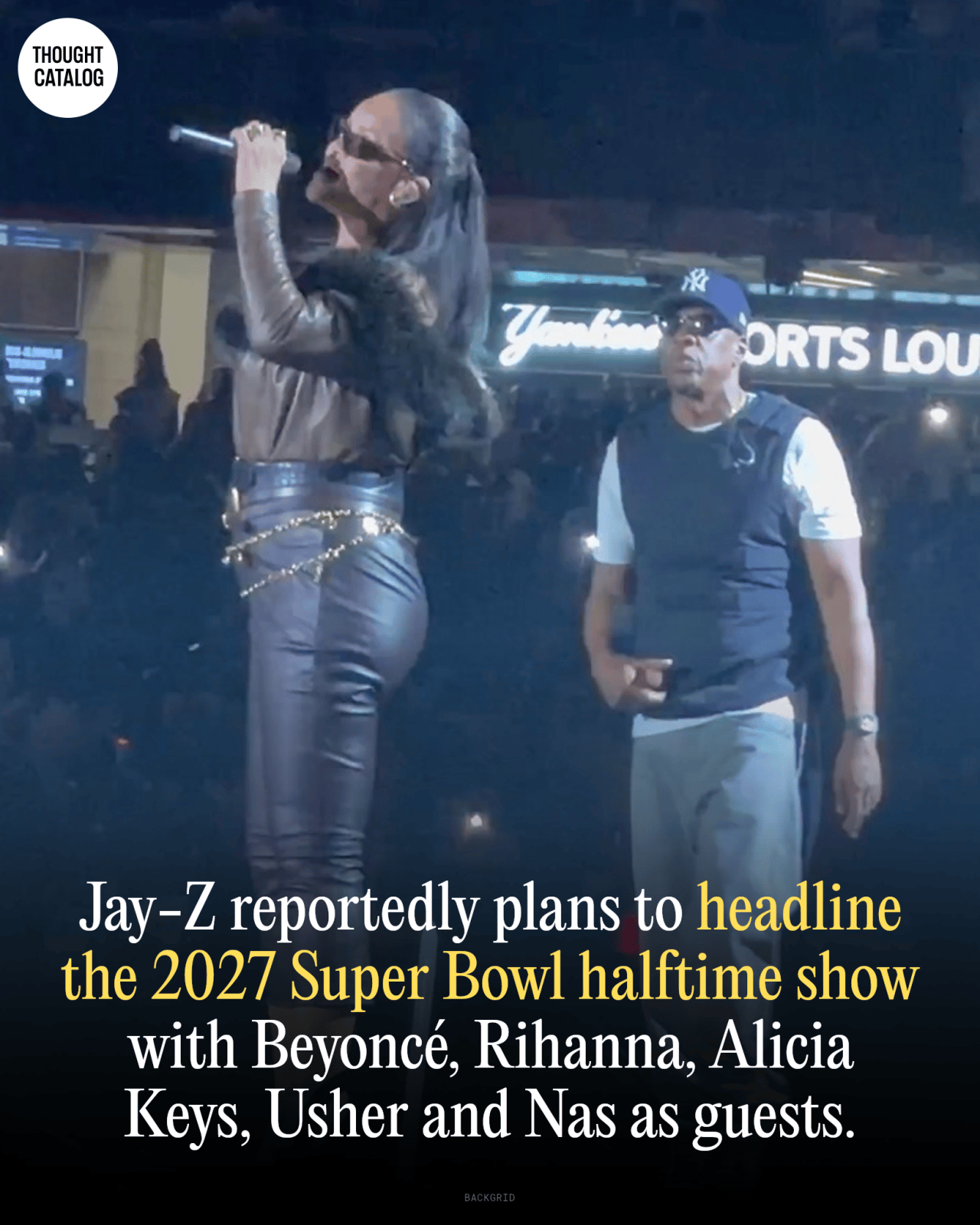 Woman in leather outfit singing on stage with a man in a Yankees cap standing behind her, outdoor venue at night.
