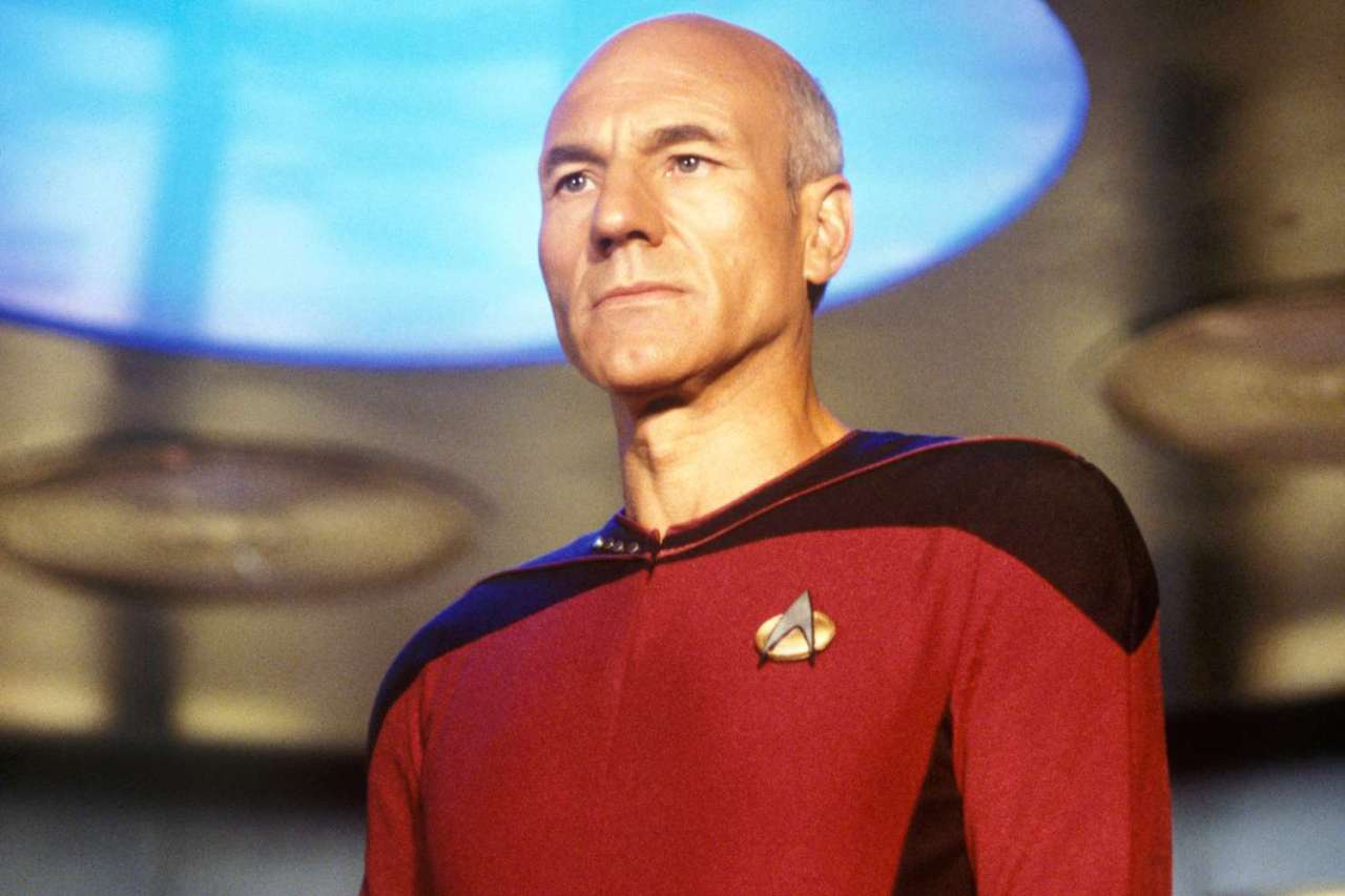 Bald man in red and black Star Trek Starfleet uniform standing on a sci-fi set with a blue viewscreen behind him.