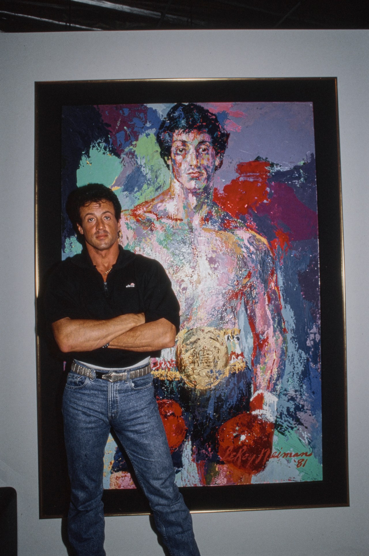 Sylvester Stallone in a black polo shirt and stonewashed jeans standing beside a LeRoy Neiman portrait of Rocky Balboa, circa 1982
