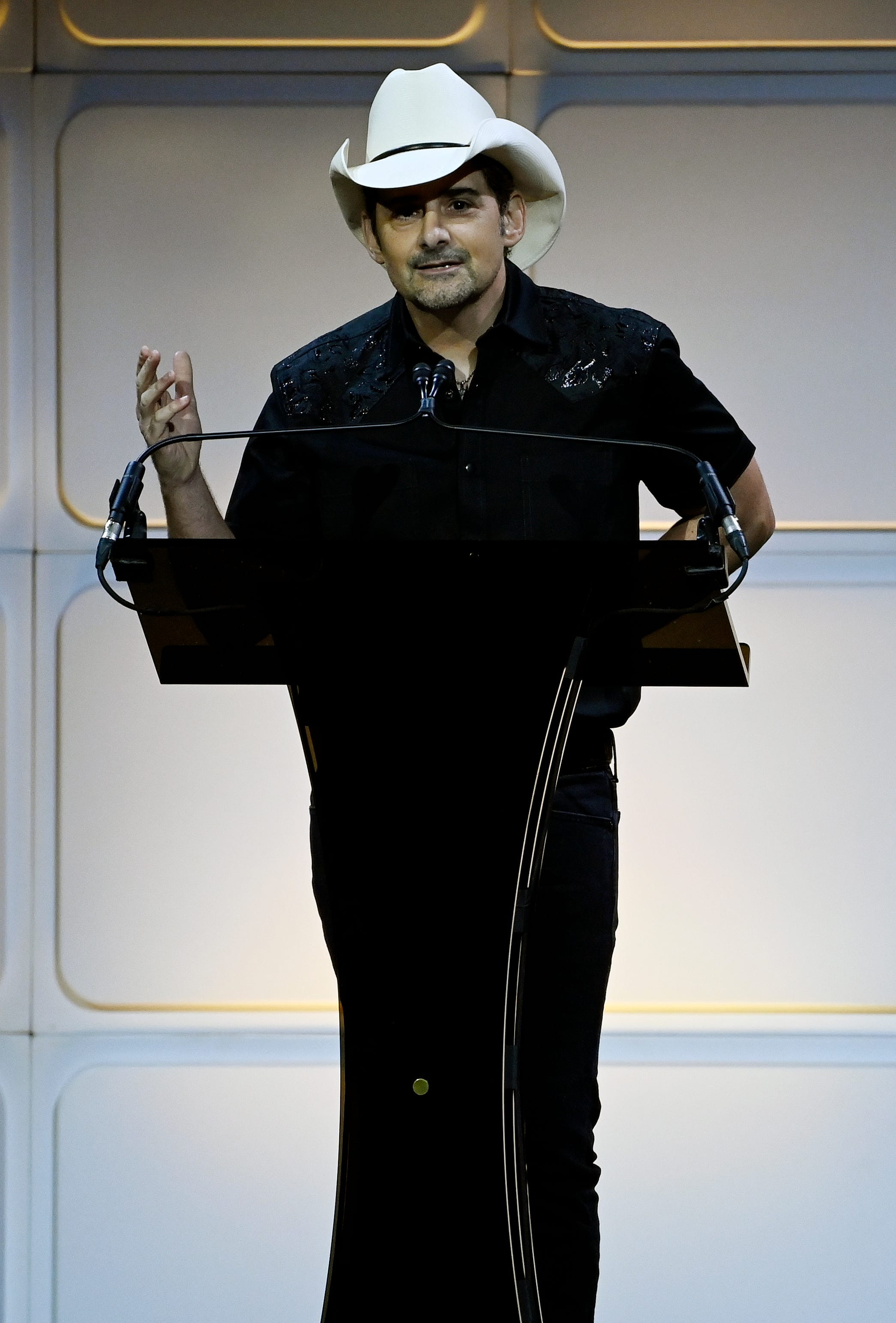 Man in a white cowboy hat and black embroidered shirt speaking at a podium on a darkened stage.